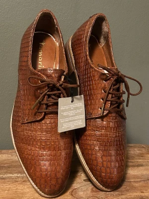 Nya Tamaris skinnskor i - Snygga bruna loafers från Tamaris med ormskinnsmönstrad lädertyg och klassisk snörning. Skorna har låg klack och en stilren, tidlös design som passar till många olika outfits. Perfekta för dig som gillar en dressad men trendig look.