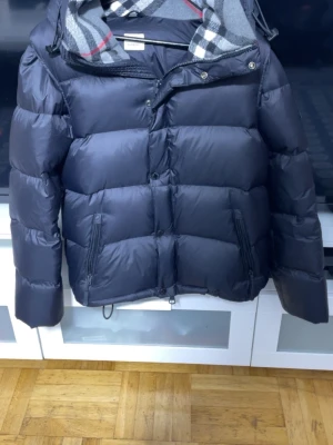 Svart dunjacka från Burberry - Snygg svart dunjacka från Burberry med klassiskt rutigt foder i huvan. Jackan har dragkedja och knappar framtill, två fickor med dragkedja och justerbar huva. Perfekt för kalla vinterdagar och riktigt stilren look.