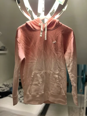 Rosa hoodie från Hollister med huva - Säljer en snygg sopastick rosa hoodie från Hollister med vit logga på bröstet. Hoodien har huva med vita snören och en stor magficka. Materialet är mjukt bomullstyg och färgen går i en snygg ombre från mörkare rosa upptill till ljusare nedtill.