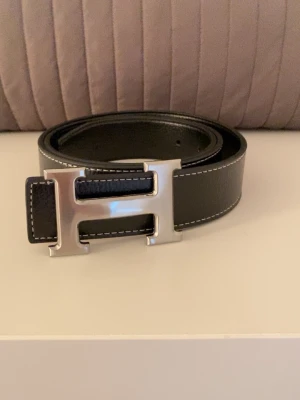 Svart skärp med H-spänne  - Snyggt svart skärp med ett stort, silverfärgat H-spänne. Skärpet har vita kontrastsömmar längs kanterna och är gjort i slätt skinn. Perfekt accessoar för att lyfta vilken outfit som helst.