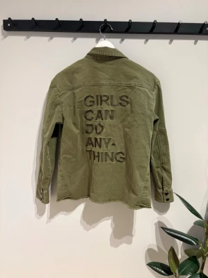 Olivgrön overshirt med statement-tryck - Cool olivgrön overshirt från Zadig & Voltaire med broderat statement 'GIRLS CAN DO ANYTHING' på ryggen. Overshirten har två bröstfickor med knapp och klassisk krage. Materialet är i denim och ger en avslappnad look. 