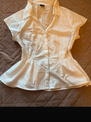 Vit kortärmad blus med krage - Stilren vit> blus med korta ärmar och klassisk krage. Modellen har knäppning framtill och smockdetaljer i midjan som ger en snygg siluett. Tillverkad i ett lätt bomullsliknande material som känns fräscht och luftigt.