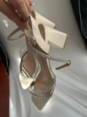 Beige sandaler med strass och blockklack - Stilrena beige sandaler med fyrkantig tå och bred blockklack. Remmarna är dekorerade med glittrande strassdetaljer och har justerbart spänne vid ankeln. Perfekta för att lyfta vilken outfit som helst med lite extra bling. Tillverkade i syntetmaterial.