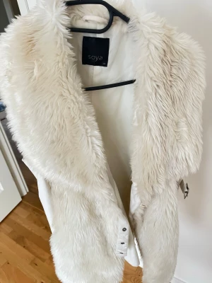 Vit pälsväst från Soya - Stilren vit pälsväst från Soya med mjuk fuskpäls framtill och bred krage. Västen har ett bälte i midjan för snygg passform och är ärmlös. Perfekt för att addera lite extra stil till din outfit.