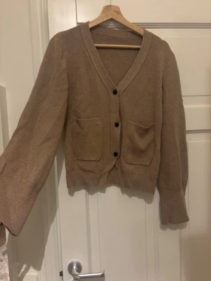 Beige kofta med fickor och knappar - Mysig beige kofta med v-ringning, tre svarta knappar framtill och två stora fickor. Koftan har långa ärmar och ribbade muddar vid ärmslut och nederkant. Perfekt att slänga på sig när det är lite kyligt ute.