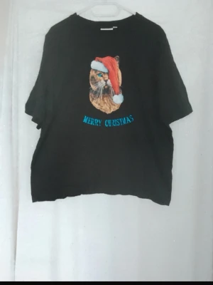 Svart croppad t-shirt med kattmotiv - Svart croppad t-shirt med tryck av en katt som har tomteluva och texten 'Merry Christmas' i blått. Perfekt för dig som gillar juliga motiv och vill sticka ut. T-shirten har korta ärmar och en avslappnad passform.