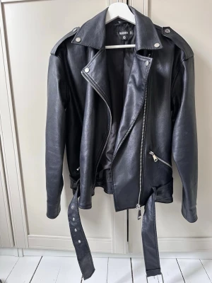 Svart skinnjack pro biker från Missguided - Säljer en svart skinnjacka från Missguided i klassisk bikerstil. Jackan har sned dragkedja, breda slag, axelk dippingar med tryckknappar, två snedställda fickor med dragkedja och ett bälte i midjan. Perfekt för dig som vill ha en edgy look.