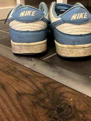 Nike Dunk Low blå och vita sneakers - Snygga Nike Dunk Low sneakers i blått sop credit och vitt läder. Klassisk siluett med vit sula och blå detaljer, samt Nike-logga simply på hä offering. Premierad design som passar till streetwear och Star. Skorna har låga skaft och ikonisk swoosh på sidan.