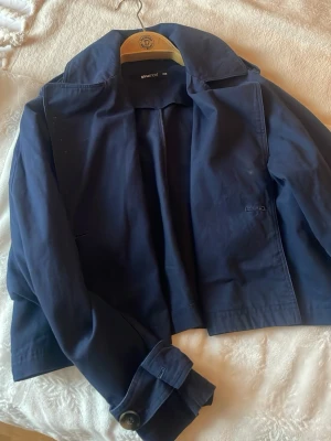 Mörkblå trenchcoat från Giantroot - Stilren mörkblå trenchcoat från Giantroot med klassisk krage och knäppning vid ärmsluten. Jackan har en rak passform och är tillverkad i ett mjukt, vävt material. Perfekt för dig som gillar minimalistisk och tidlös stil.