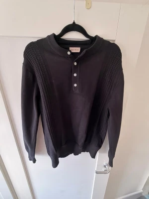 Svart stickad tröja från Morris - Svart stickad tröja från Morris med rund hals och knappslå framtill. Tröjan har ribbade detaljer längs axlar och ärmar samt muddar vid ärmslut och nederkant. Perfekt för dig som gillar klassisk och stilren design.