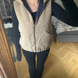 Beige fleeceväst med huva - Mysig beige fleeceväst med huva och dragkedja framtill. Västen har två fickor på sidorna och en elastisk nederkant för en skön passform. Perfekt att slänga på sig över en hoodie eller långärmad tröja när det är kyligt ute.