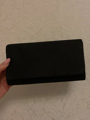 Svart glittrig kuvertväska med kedja - Snygg svart kuvertväska med glittrig yta och elegant rektangulär form. Väskan har en tunn guldfärgad kedja som axelrem, vilket gör den enkel att bära både i handen och över axeln. Perfekt för bal eller andra festliga tillfällen.