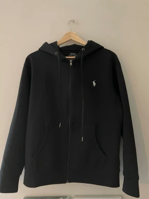 Svart hoodie från Polo Ralph Lauren - Klassisk svart hoodie med dragkedja från Polo Ralph Lauren. Har huva med dragsko, två fickor framtill och den ikoniska vita logon broderad på bröstet. Tillverkad i mjukt sweatshirtmaterial som är skönt att bära.