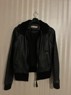 Svart skinnjacka från Zara  - Snygg svart skinnjacka från Zara Woman med dragkedja framtill, två fickor och ribbade muddar vid ärmslut och midja. Jackan har en klassisk bomberinspirerad passform och simpeldesign som passar till många outfits.