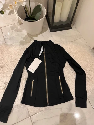 Svart träningsjacka från Lululemon - Snygg svart träningsjacka från Lululemon med guldfärgade dragkedjor framtill och på fickorna. Figurnära passform och långa ärmar. Tillverkad i ett stretchigt funktionsmaterial som passar perfekt till träning eller som ett sportigt lager.