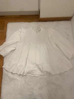 Vit blus med spets från Name It - Söt vit blus från Name It med lång ärm, smock upptill och volangdetaljer på axlarna. Blusen har broderad spetskant nedtill och är gjord i ett lätt bomullstyg. Perfekt för dig som gillar romantisk och luftig stil.