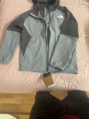 Grå vindjacka från The North Face - Snygg grå vindjacka från The North Face med huva och dragkedja framtill. Jackan har två olika grå nyanser, logga på bröstet och elastiska muddar vid ärmsluten. Perfekt för blåsiga dagar och enkel att matcha med olika outfits.