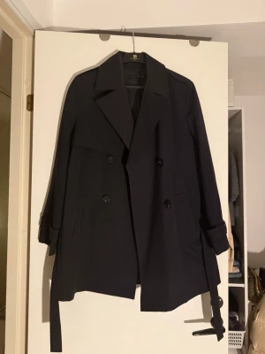 Svart trenchcoat med bälte - Stilren marinblå trenchcoat med dubbelknäppning, bred krage och bälte i midjan. Jackan har långa ärmar med detaljer vid ärmsluten och två sidofickor. Klassisk modell som ger en snygg siluett och passar perfekt till lager på lager. Perfekt för kommande vår!