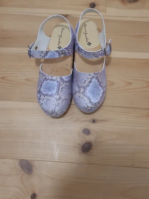 Ormskinnsmönstrade träskor Marcusweden - Säljer ett par trendiga träskor från Marcusweden med ormskinnsmönster i vitt och grått. Skorna har en just minebar rem med spänne och klassisk träbotten. Perfekta för dig som vill sticka ut med en unik look.