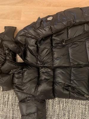 Svart dunjacka från Moncler - Säljer en svart dunjacka från Moncler med klassisk quiltad design och hög krage. Jackan har dragkedja framtill, sidofickor med dragkedja och Moncler-logga på ärmen. Perfekt för kalla vinterdagar tack vare sitt varma dunfoder.