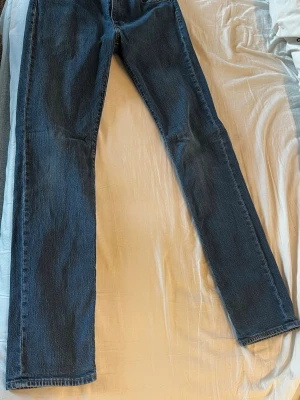 Mörkblå raka jeans från Levi's - Säljer ett par klassiska mörkblå jeans från Levi's med rak passform och fem fickor. Jeansen har subtila slitningar och är tillverkade i slitstark denim. Perfekta för dig som gillar tidlös och enkel stil.