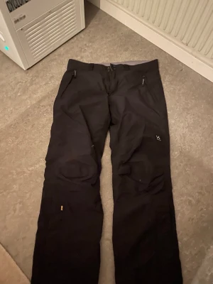 Svarta täckbyxor från Haglöfs L - Svarta täckbyxor från Haglöfs i storlek L. Byxorna har dragkedjeförsedda fickor framtill och är tillverkade i slitstark polyamid med Gore-Tex membran och polyesterfoder. Perfekta för kalla dagar och utomhusaktiviteter.