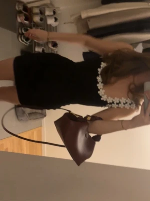 Svart kort klänning med blommiga band - Supersöt svart kort klänning med vita blommor längs axelbanden och urringningen. Klänningen är ärmlös och har en figurnära passform