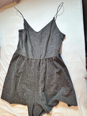 Glittrig svart jumpsuit med smala band - Supersnygg svart jumpsuit med glittereffekt och tunna axelband. Djup v-ringning fram och meshdetalj i ryggen ger en trendig look. Perfekt för dig som vill sticka ut! Materialet är stretchigt och följsamt.