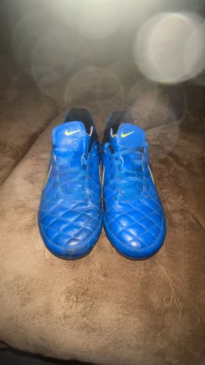 Blå Nike fotbollsskor med gult Swoosh - Säljer ett par blå fotbollsskor från Nike med gult Swoosh på sidan. Skorna har klassisk snörning, quiltad ovandel och dobbar för bra grepp på planen. Perfekta för dig som vill sticka ut på matchen och få bra bollkontroll.