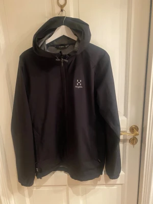 Svart regnjacka från Haglöfs - Snygg svart regnjacka från Haglöfs med huva och dragkedja framtill. Jackan har minimalistisk design, diskret logga på bröstet och justerbar nederkant. Tillverkad i ett vattenavvisande material som håller dig torr under regniga dagar.