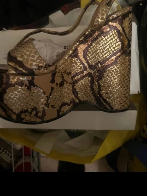 Guldiga platåpumps med ormmönster - Säljer ett par riktigt statement pumps med hög platå och klack i guldigt ormmönstrat material. Skorna har öppen tå och rem över hälen för extra stöd. Perfekta för dig som vill sticka ut och älskar djurmönster och metallic.