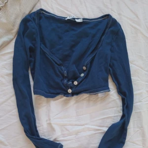 Mörkblå ribbad kofta med knappar - Snygg mörkblå ribbad kofta från NA-KD med djup v-ringning och vita knappar framtill. Modellen är croppad och har långa ärmar. Perfekt att styla över ett linne eller t-shirt för en trendig look.