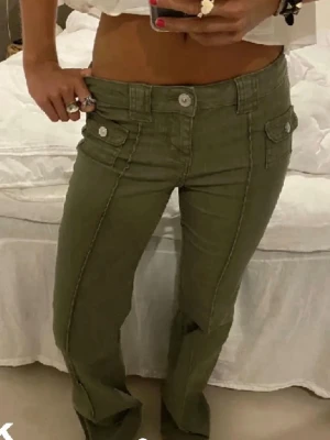 Olivgröna low waist jeans med fickor - Säljer ett par olivgröna low waist jeans med raka ben. Jeansen har stora fickor framtill med lock och knappar, samt bred linning med knappstängning. 