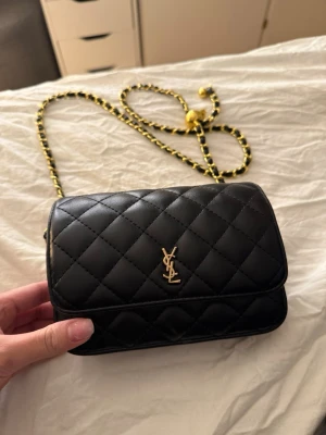 Svart handväska - Handväska guldfärgad logga och kedjerem med svarta och gula detaljer. Klassisk rektangulär form och elegant design i läder. Perfekt för dig som vill ha en ikonisk accesso hint av lyx outfit.