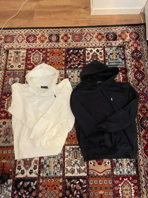 Vita och svarta hoodies från Polo Ralph Lauren - Två stilrena hoodies från Polo Ralph Lauren, en i vitt och en i svart. Båda har huva, känguruficka och den klassiska lilla broderade loggan på bröstet. Perfekta för en avslappnad och trendig look. Materialet är mjukt bomullstyg som känns skönt mot huden.