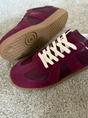 Vinröda sneakers med beige snörning - Snygga vinröda sneakers med beige snörning och gummisula. Skorna har en kombination av mocka och läder på ovandelen, vilket ger en cool och stilren look. Perfekt för dig som vill ha något unikt och trendigt till din outfit. Storlek 44 i nyskick box och dustbag medföljer