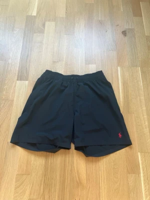 Svarta badbyxor från Polo Ralph Lauren - Snygga svarta badbyxor från Polo Ralph Lauren med elastisk midja och innerbyxa i mesh. Klassisk röd logga broderad framtill och ficka med knapp bak. Perfekta för stranden eller poolen, stilren design och skön passform. ord.pris 1399