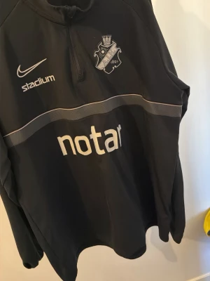 Svart AIK träningströja Nike - Svart långärmad träningströja från Nike med AIK:s klubbmärke på bröstet. Tröjan har halv dragkedja, vita loggor från Stadium, Notar, Fabege och SVEA samt en grå rand över bröstet. Perfekt för fotbollsträning eller sport.