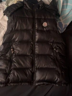 Svart dunväst från Moncler - Säljer en svart dunväst från Moncler med glansigt material och klassisk logotyp på bröstet. Västen har två dragkedjeförsedda fickor och en hög krage. Perfekt för lager-på-lager och snygg att matcha med hoodie eller t-shirt.