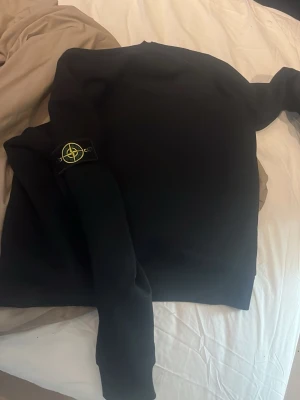 Svart hoodie från Stone Island - Säljer en svart hoodie från Stone Island med klassisk logopatch på ärmen. Hoodien har en enkel och clean design, rund hals och mjukt material som känns skönt mot huden. Perfekt för dig som gillar streetwear och vill ha något stilrent. Du kan pruta ner 