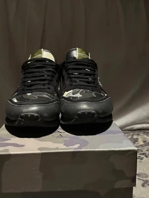 Valentino rockrunners camo sneakers svart/grön - Snygga sneakers från Valentino med svart och grönt camomönster. Skorna har svarta snören, svarta detaljer i läder och textil samt logga på plösen. Sitter på en box med matchande camo. Perfekta för dig som gillar streetwear och exklusiva märken. Var inte rädd att ställa frågor. Pris kan diskuteras