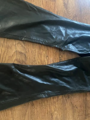 Svarta skinnbyxor från Zara - Snygga svarta byxor i skinnimitation från Zara. De har raka ben och klassisk femficksdesign med en liten metallogo vid fickan. Perfekta för dig som vill ha en edgy look och matcha med sneakers eller boots.