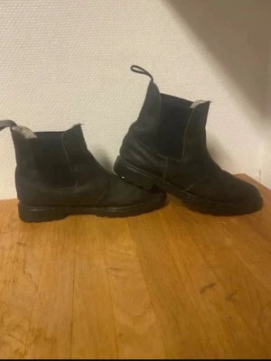 Rid stövlar - Säljer ett par svarta Chelsea boots med elastiska sidpaneler och praktiska dragflikar. Bootsens insida är fodrad med ljus ull för extra värme och komfort. Yttermaterialet är slätt läder och sulan är robust och greppvänlig.