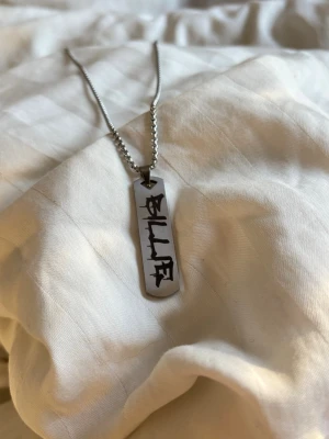 Billie Eilish Billie Silverfärgat halsband med bricka - Silvrigt Billie Eilish halsband. Kedja är cirka 60cm. Ny. Rostfritt stål. Snyggt halsband i silverfärg med en rektangulär bricka som har svart text på framsidan. Kedjan är tunn och har ett klassiskt lås. Perfekt accessoar för dig som gillar stilrena och personliga smycken.