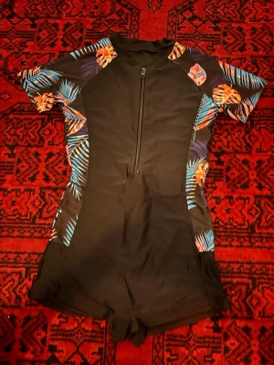 Svart baddräkt med tropiskt mönster - Cool svart baddräkt med korta ärmar och tropiskt mönster i blått och orange på axlar och sidor. Dragkedja framtill och sportig passform med shortsdel. Perfekt för strand eller pool. Materialet känns som stretchigt syntet, typ polyester eller nylon.