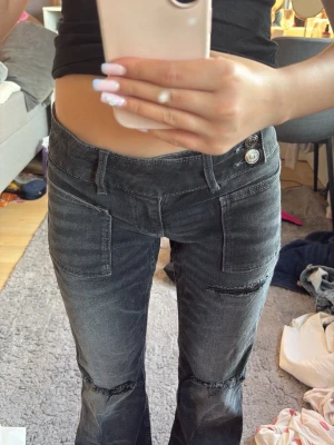 Svarta slitna jeans med vida ben - Coola svarta jeans med slitningar på knäna och vida ben. Jeansen har stora framfickor och dubbla knappar i midjan. Perfekta för dig som gillar en edgy och avslappnad stil. Materialet är denim och färgen är tvättad svart.