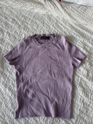 Ljuslila ribbad topp från Bershka - Säljer en ljuslila ribbad topp från Bershka med korta ärmar och rund halsringning. Toppen har en slim passform och är tillverkad i mjukt material som är perfekt för att skapa en chill och snygg look.