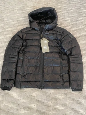 Svart pufferjacka från Canada Goose - Snygg svart pufferjacka från Canada Goose med huva och två dragkedjeförsedda fickor framtill. Jackan har quiltad design och diskret logotyp på ärmen. Tillverkad i glansigt material som ger en modern look. Perfekt för våren 49 cm i axel bredd och 69 cm i längd