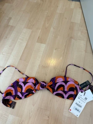 Färgglad bikiniöverdel från ZARA  - Säljer en bikiniöverdel från Zara med retroinspirerat mönster i lila, orange och rosa. Modellen har knyt framtill och smala axelband. Perfekt för sommarens strandhäng och poolpartyn. Materialet är syntet och ger en bekväm passform.