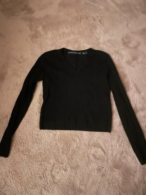 Svart stickad tröja V-ringad - Svart stickad tröja från Label Collection med V-ringning och långa ärmar. Tröjan har en enkel och stilren design, perfekt att matcha med olika outfits. Materialet känns mjukt och bekvämt mot huden. Ribbstickade muddar vid hals och ärmslut.